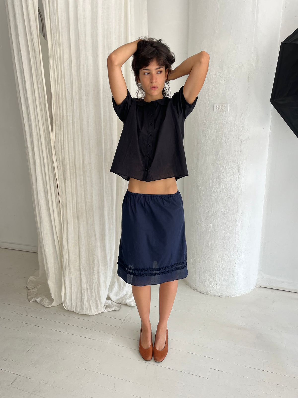 A woman stands against a white curtain with arms raised, wearing the Daytime Skirt - Navy by Deiji Studios, a cotton voile midi skirt with double frill detailing at the hem, styled with the Petal Top in Black.
