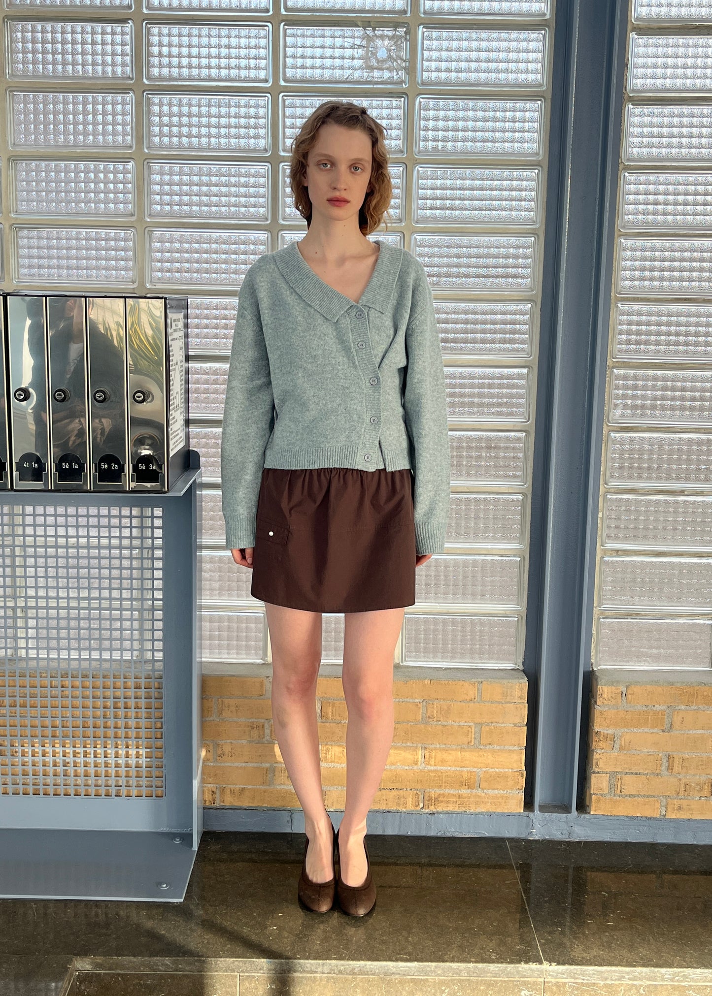 A person with short, wavy hair stands indoors, wearing a light gray buttoned cardigan and the Pocket Skirt - Sepia. Facing forward with a neutral expression before glass brick walls and a shelf with binders, they evoke Deiji Studios style.
