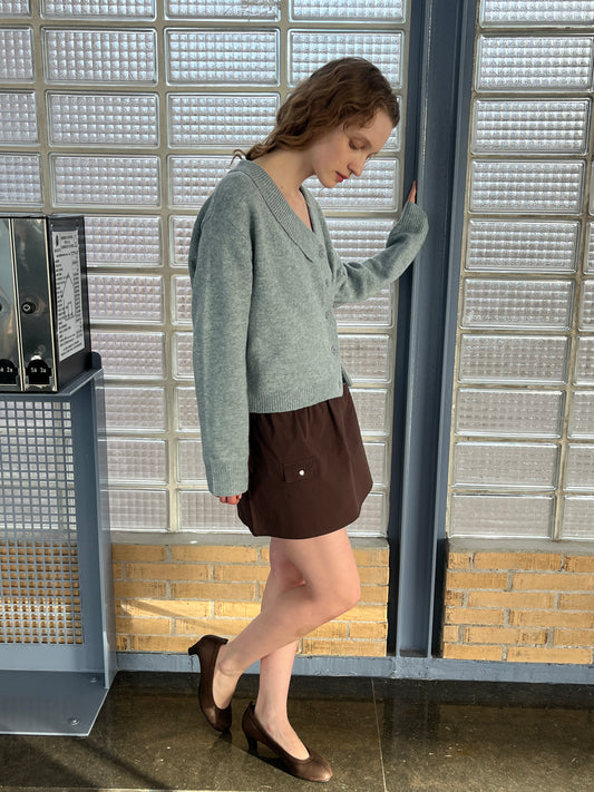 A woman with wavy hair stands indoors against a blue metal beam, wearing a Deiji Studios gray cardigan, the Pocket Skirt - Sepia, and brown heels. The scene features glass blocks and brick in the background.