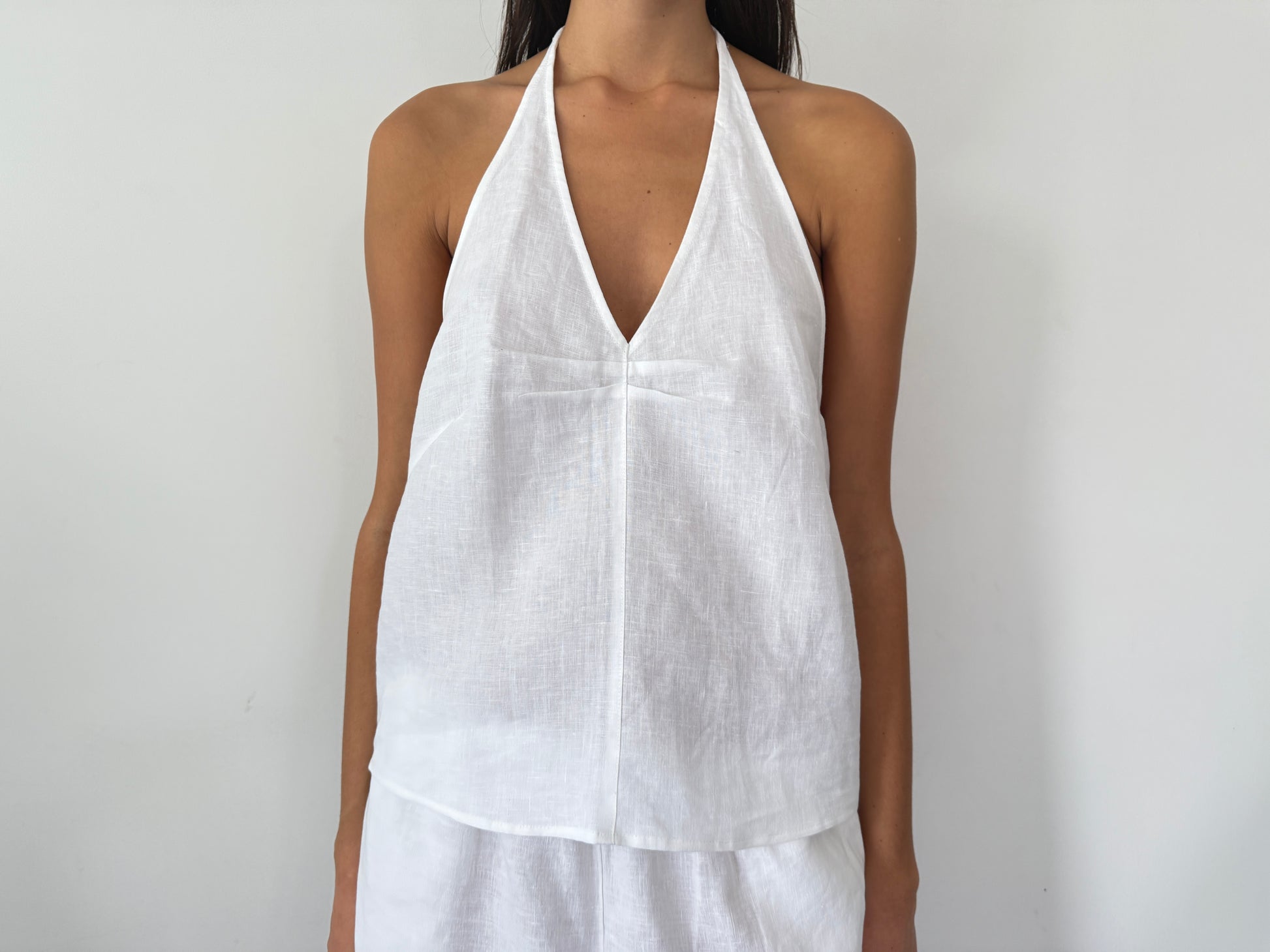 A person wearing the Linen Pinched Halter Top - White, a sleeveless French linen halter with a V neckline, stands against a plain white background. Only the upper body is visible.
