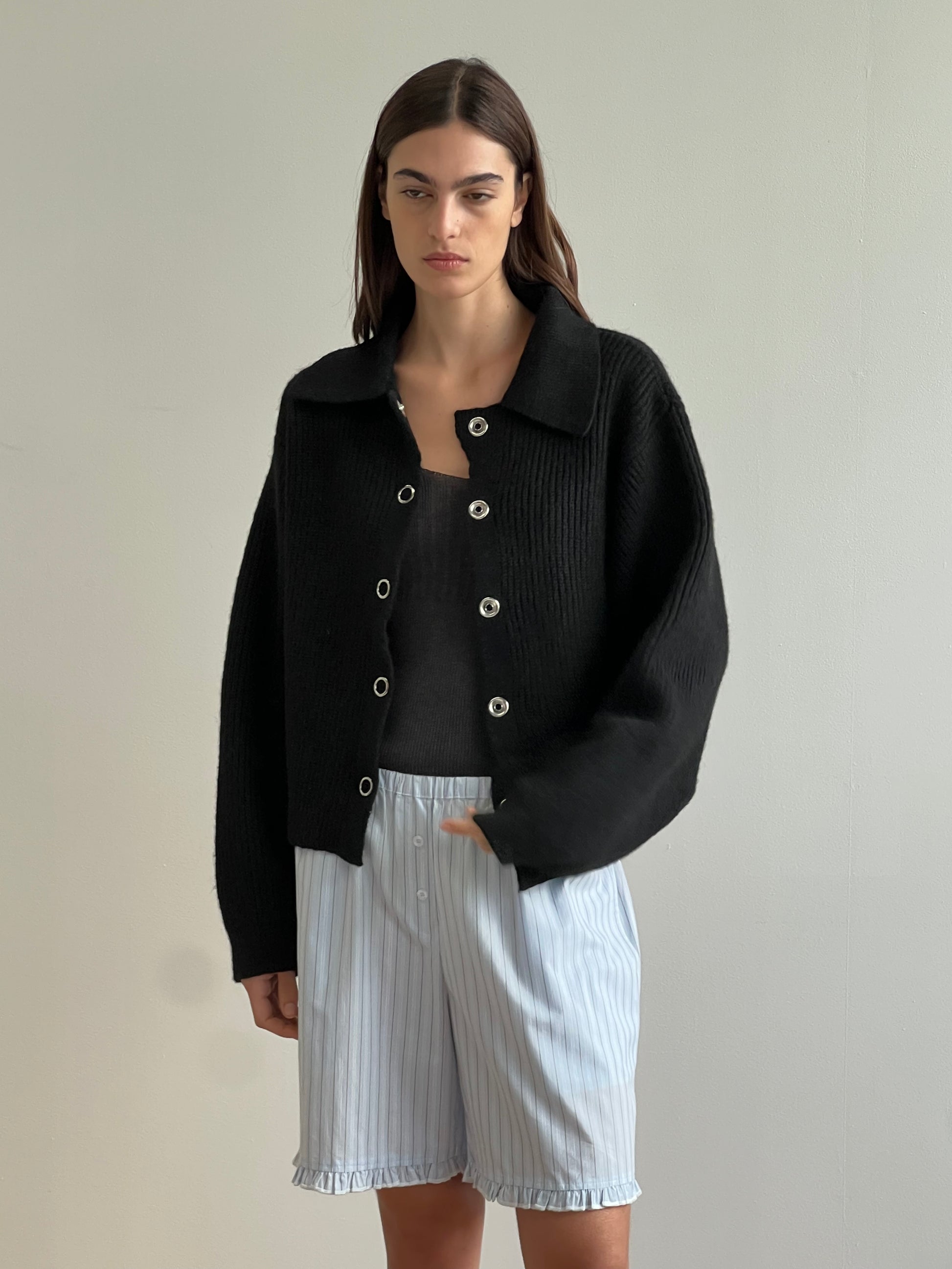 A woman with straight brown hair wears the sustainable Stud Cardi Knit - Black in an oversized fit over a black top and loose, light blue pinstriped ruffle-hem shorts, standing against a plain light-colored wall.