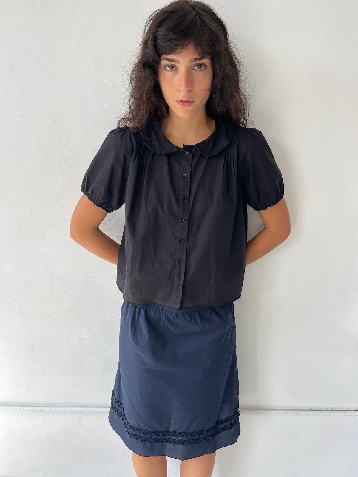 A woman with dark wavy hair stands against a plain white wall, wearing the Petal Top - Black by Deiji Studios, a cotton voile top with a peter pan collar and puffed short sleeves, styled with the Daytime Skirt in Navy.