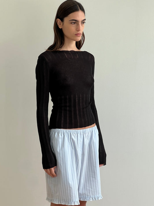 A woman with long dark hair stands against a plain light wall, wearing the Flounce Short - Mineral Stripe and a fitted, sheer black long-sleeve top.