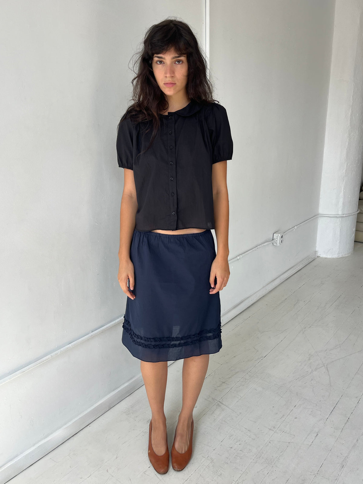 A woman with dark wavy hair stands against a white wall, wearing the Daytime Skirt - Navy by Deiji Studios, a cotton voile midi skirt with a straight silhouette and double frill detailing at the hem, styled with the Petal Top in Black.