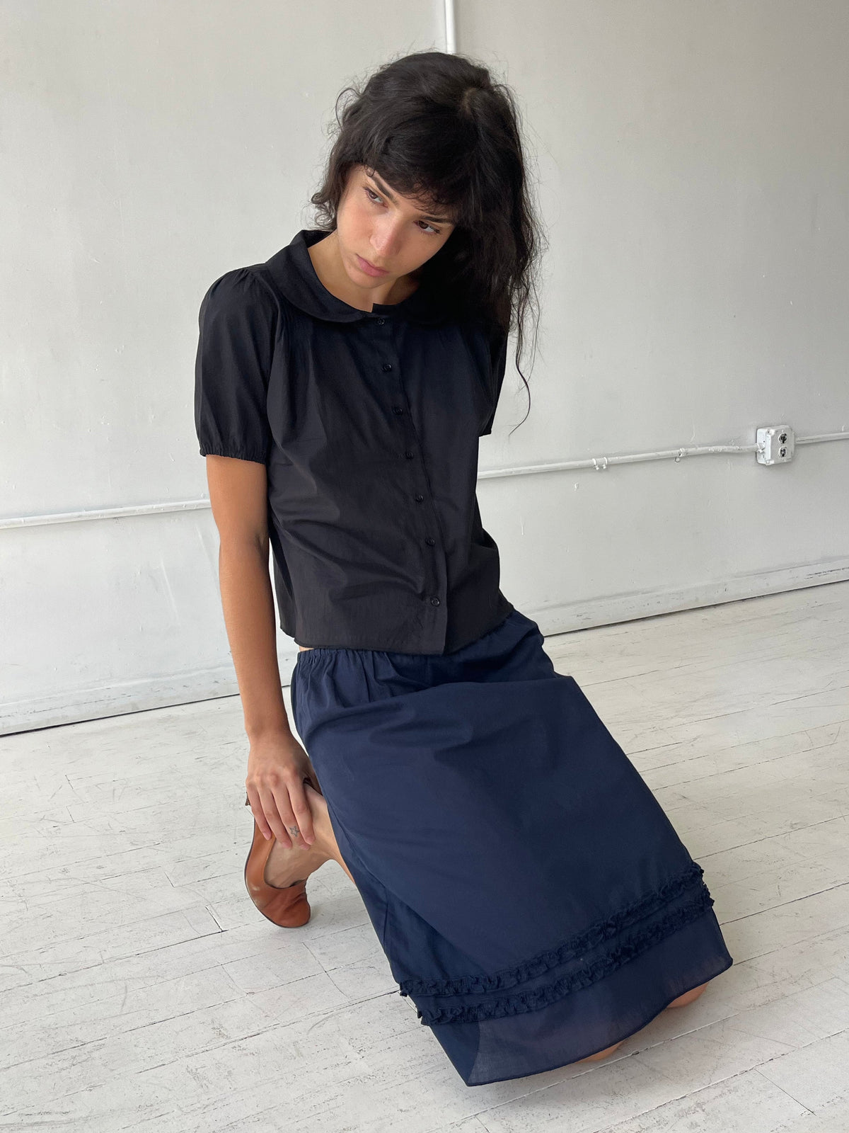 A woman kneels on a white studio floor, wearing the Daytime Skirt - Navy by Deiji Studios, a cotton voile midi skirt with double frill detailing at the hem, styled with the Petal Top in Black.