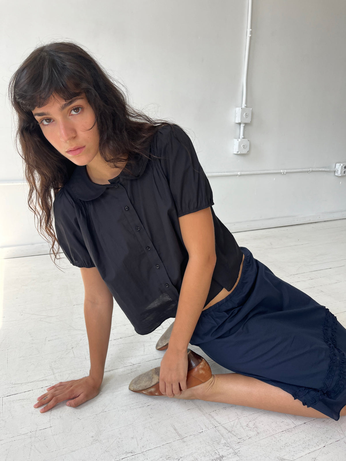 A woman sits on a white studio floor, wearing the Petal Top - Black by Deiji Studios, a cotton voile top with a peter pan collar and puffed short sleeves, styled with the Daytime Skirt in Navy.