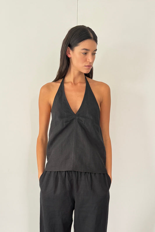 A woman stands indoors against white double doors, wearing the Linen Pinched Halter Top in black with loose black pants and sandals. Clothes hang on a rack to her right as she looks to her left.