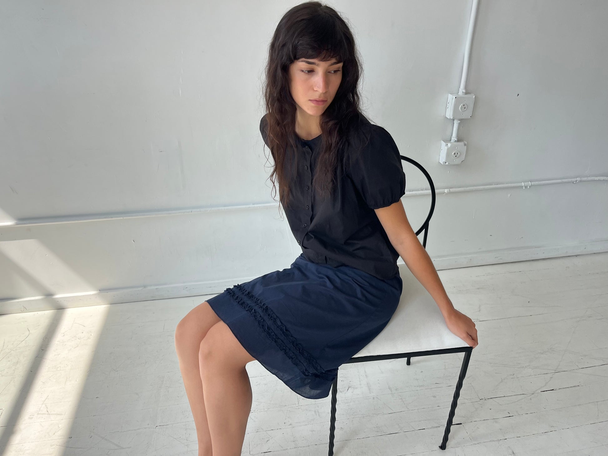 A woman sits on a white bench in a studio, wearing the Daytime Skirt - Navy by Deiji Studios, a cotton voile midi skirt with double frill detailing at the hem, styled with the Petal Top in Black.