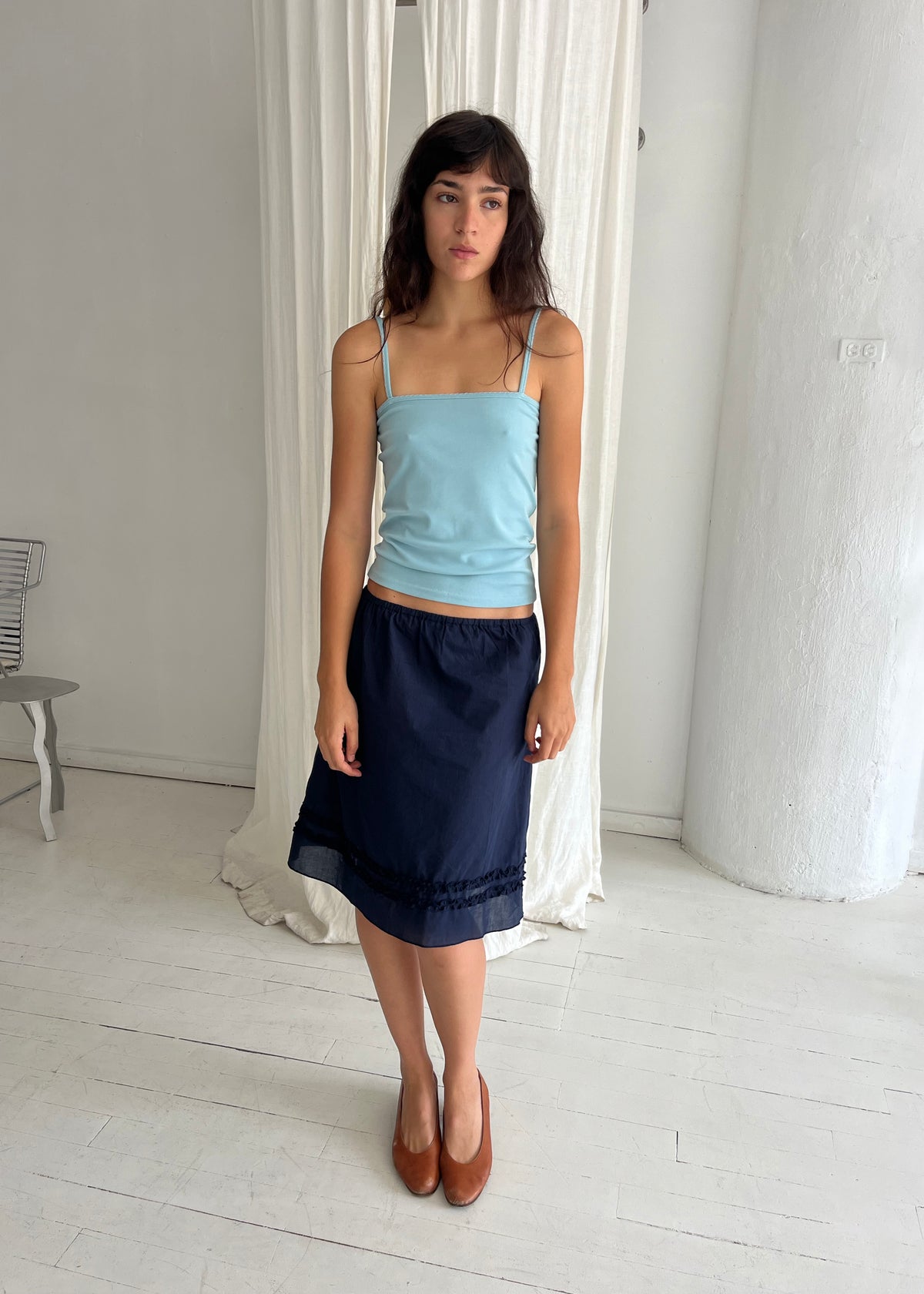 A woman stands against a white curtain, wearing the Daytime Skirt - Navy by Deiji Studios, a cotton voile midi skirt with a straight silhouette and double frill detailing at the hem, styled with the Scallop Tank in Sky.