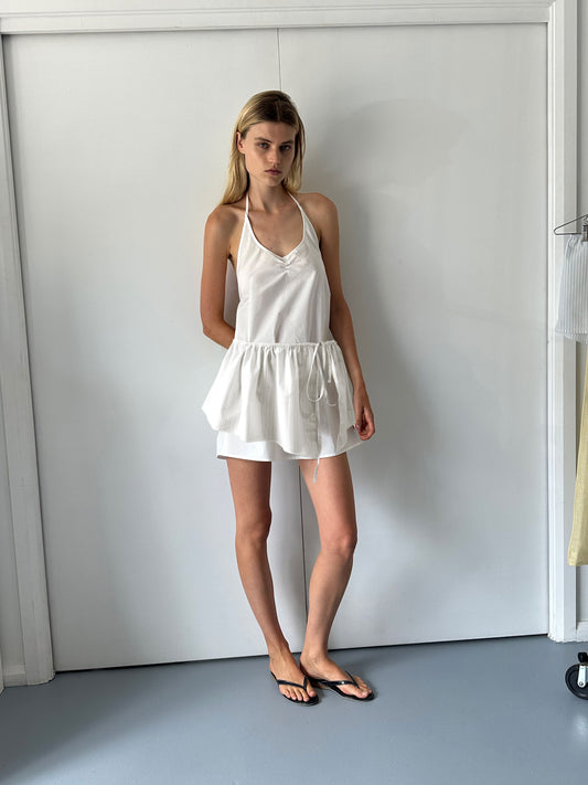 Against a white wall, a woman with straight blonde hair and a neutral expression wears white organic cotton shorts, a sleeveless halter top with ruffled hem, black flip-flops, and accessorizes with the Napkin Belt - Pacific Stripe.
