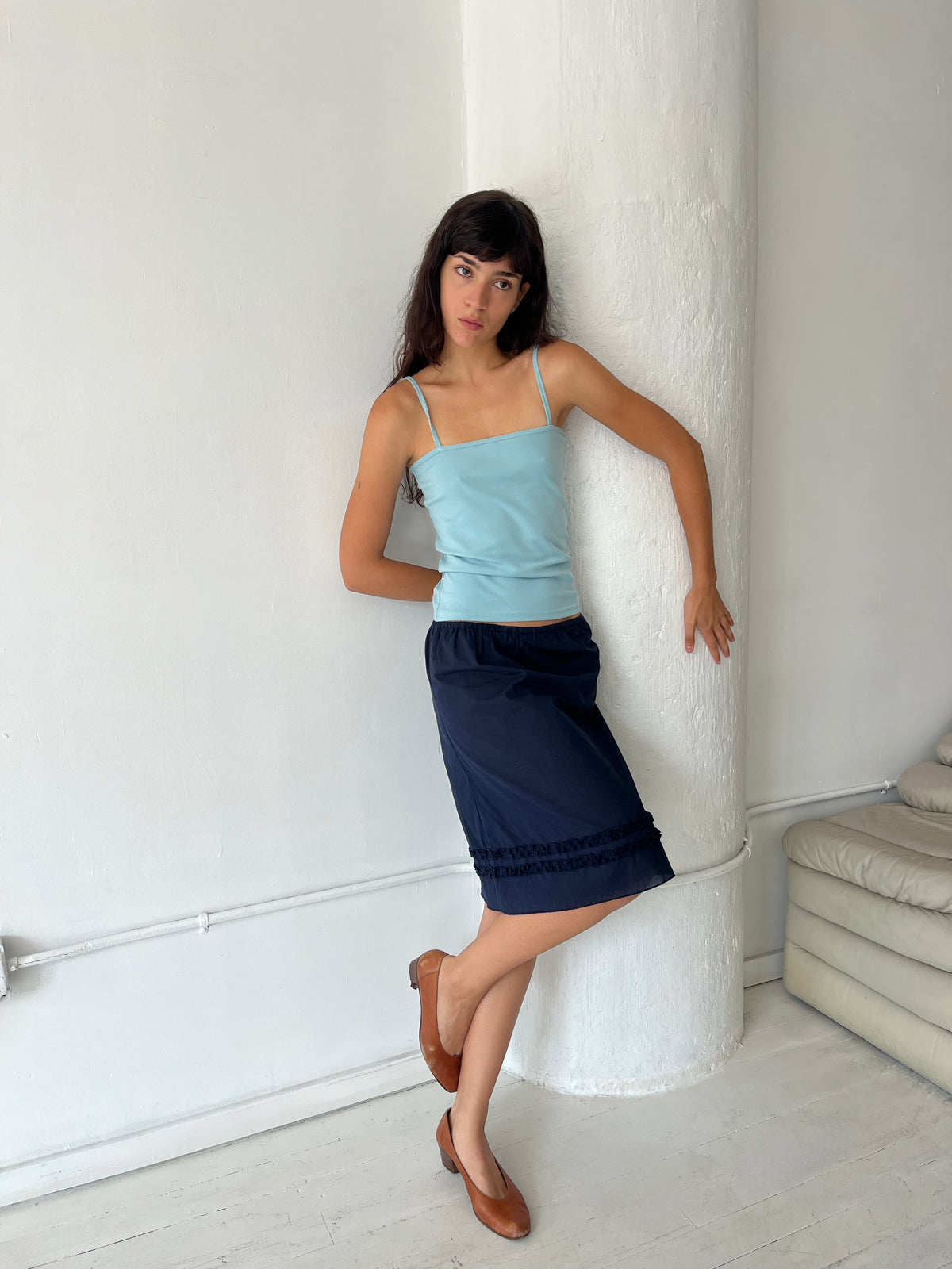 A woman leans against a white column, wearing the Daytime Skirt - Navy by Deiji Studios, a cotton voile midi skirt with double frill detailing at the hem, styled with the Scallop Tank in Sky and brown flats.