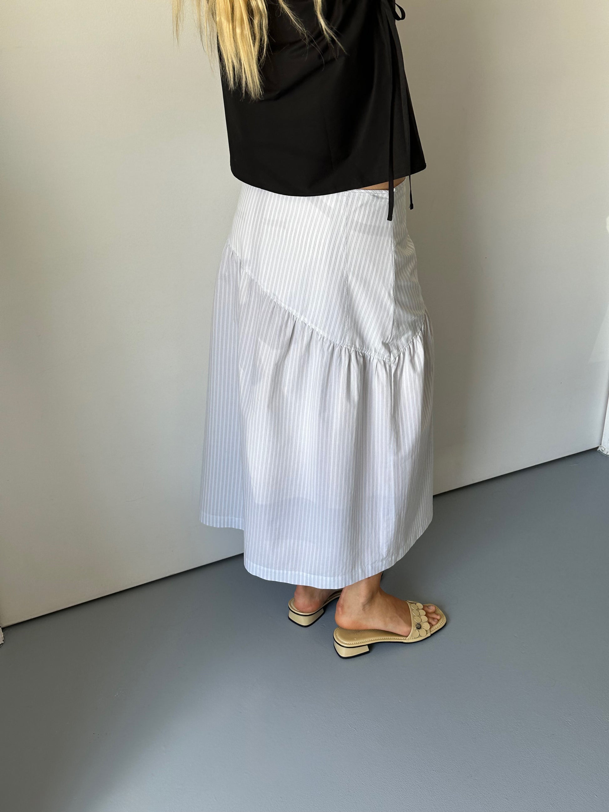 A person with long blonde hair wears a black top, the 0 Angle Skirt - Sage Stripe, and beige open-toed sandals, standing indoors on a light grey floor against a white wall.