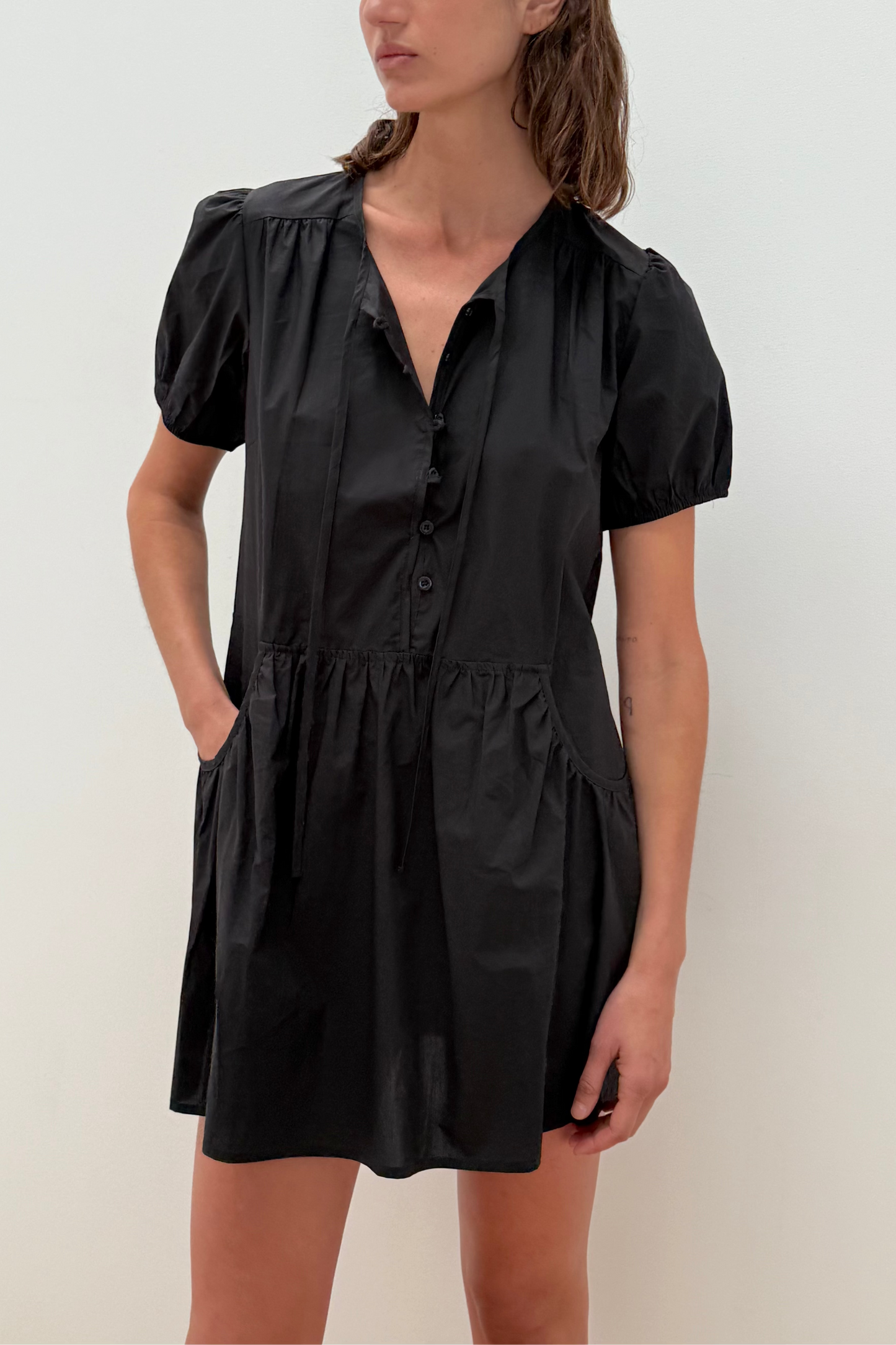 A woman wears the Pocket Mini Dress - Black by Deiji Studios, made from organic cotton with short puff sleeves, a button-up front, and pockets, standing against a plain white background.