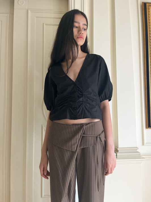 A woman with long dark hair wears a black Draw Top—organic cotton, short-sleeved, v neckline—paired with high-waisted brown pinstriped pants. She stands indoors against an off-white wall with molding, looking to the side.