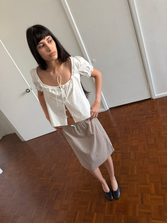 A woman with dark hair and a fringe stands in a room wearing an open white frill top and the Basic Skirt - Grey by Deiji Studios, a grey cotton voile midi skirt, with black flats.