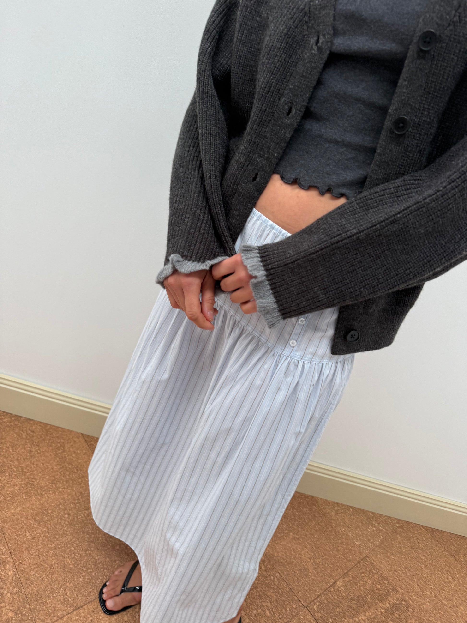 A person stands indoors on a brown floor, hands clasped, wearing a dark gray sweater over a matching top and the Button Skirt - Mineral Stripe—a long white organic cotton skirt with thin blue stripes. Their face is not visible.