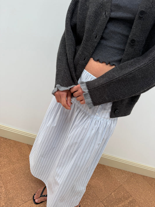 A person stands indoors on a brown floor, hands clasped, wearing a dark gray sweater over a matching top and the Button Skirt - Mineral Stripe—a long white organic cotton skirt with thin blue stripes. Their face is not visible.