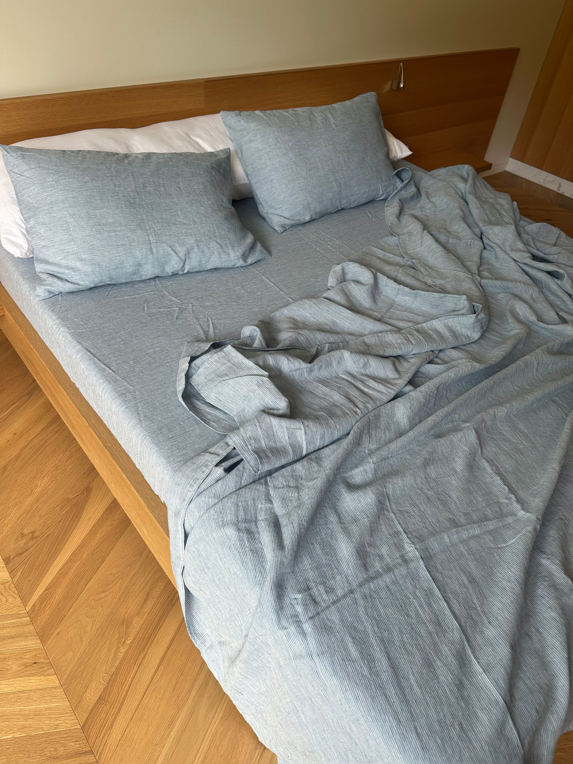 A wooden bed with Sheets - Blue Stripe, two pillows, and a slightly messy sheet partially hanging off the side, in a room with wooden flooring.
