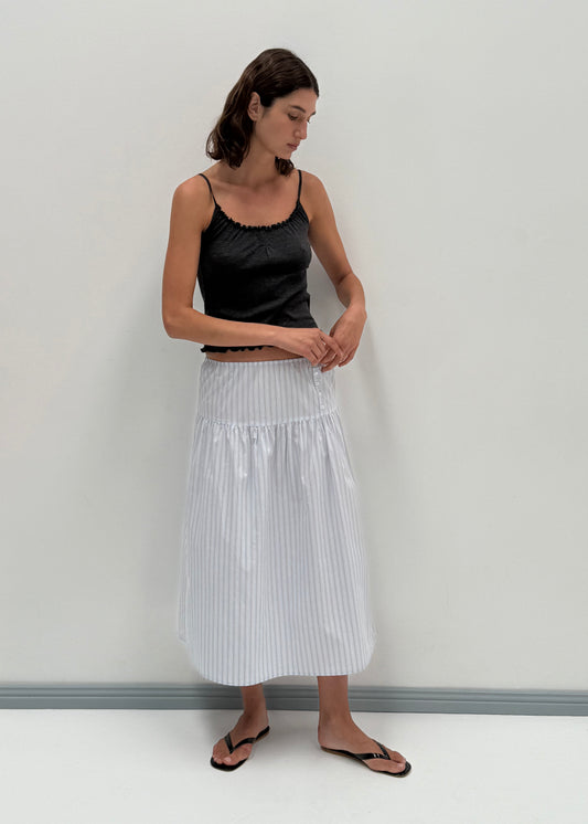 A woman stands against a plain white wall, wearing a black sleeveless top, black flip-flops, and the Button Skirt - Mineral Stripe by Deiji Studios in white and light blue stripes. She looks down with her hands lightly clasped.