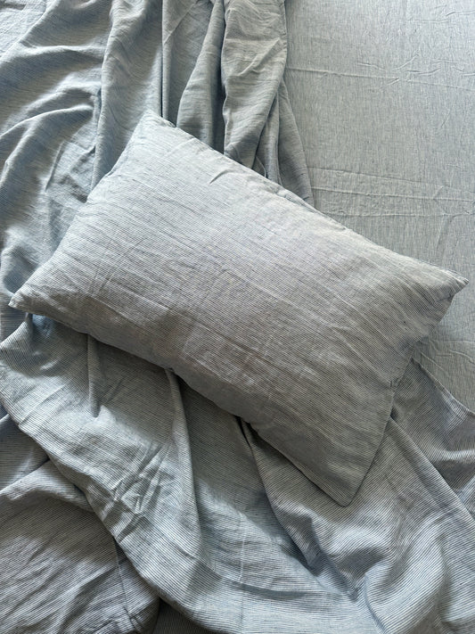 A light gray pillow rests on a bed covered with Sheets - Blue Stripe by Deiji Studios and a crumpled blanket. The fine-textured linen creates a calm, tidy atmosphere.
