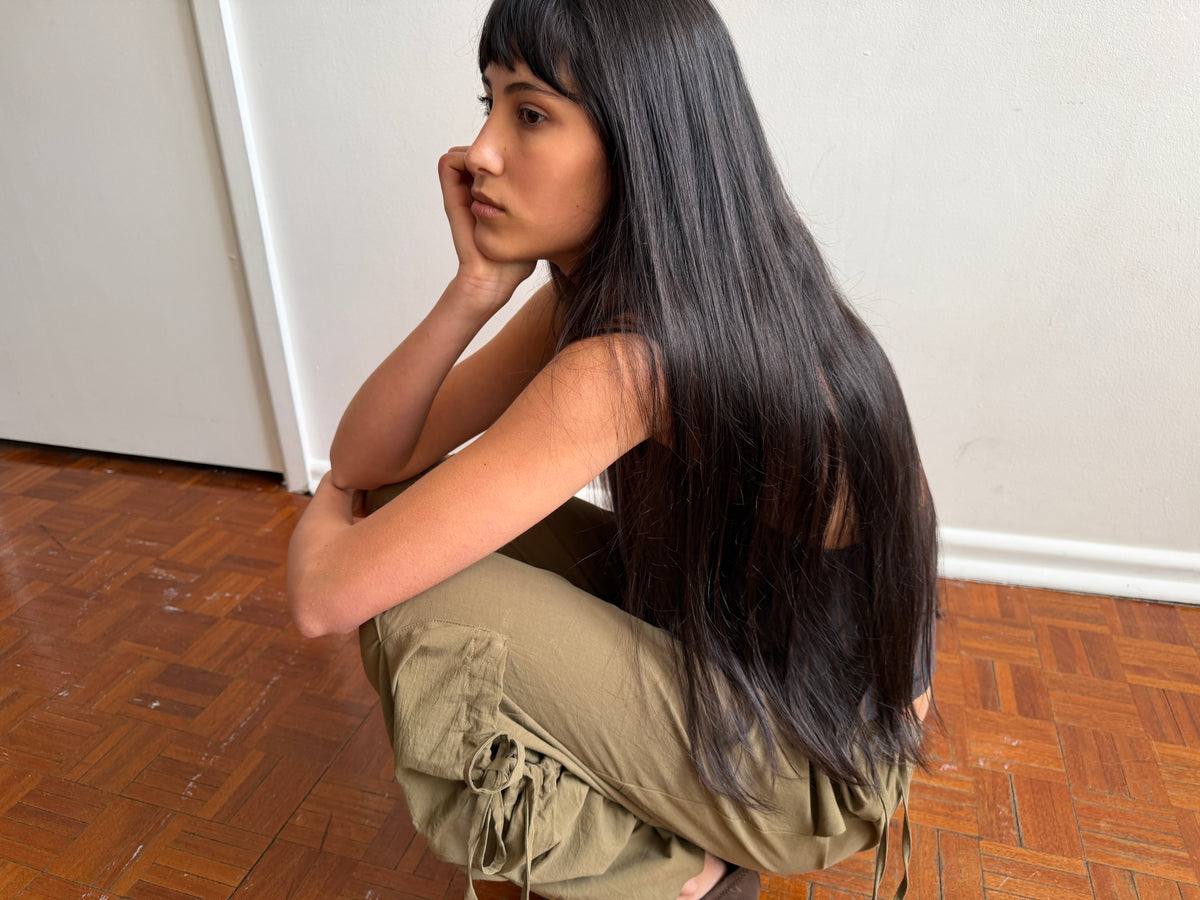 A woman with dark hair and a fringe crouches against a wall in a room wearing the Drawcord Pocket Pant - Khaki by Deiji Studios, khaki wide-leg trousers with a drawcord tie, resting her chin on her hand.