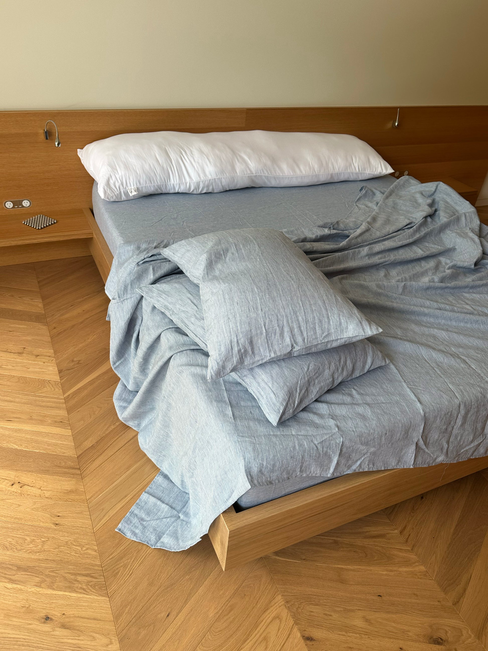 An unmade bed featuring Sheets - Blue Stripe sits against a wood-paneled wall, with two pillows and rumpled bedding draping over the side, partially revealing a herringbone wood floor.