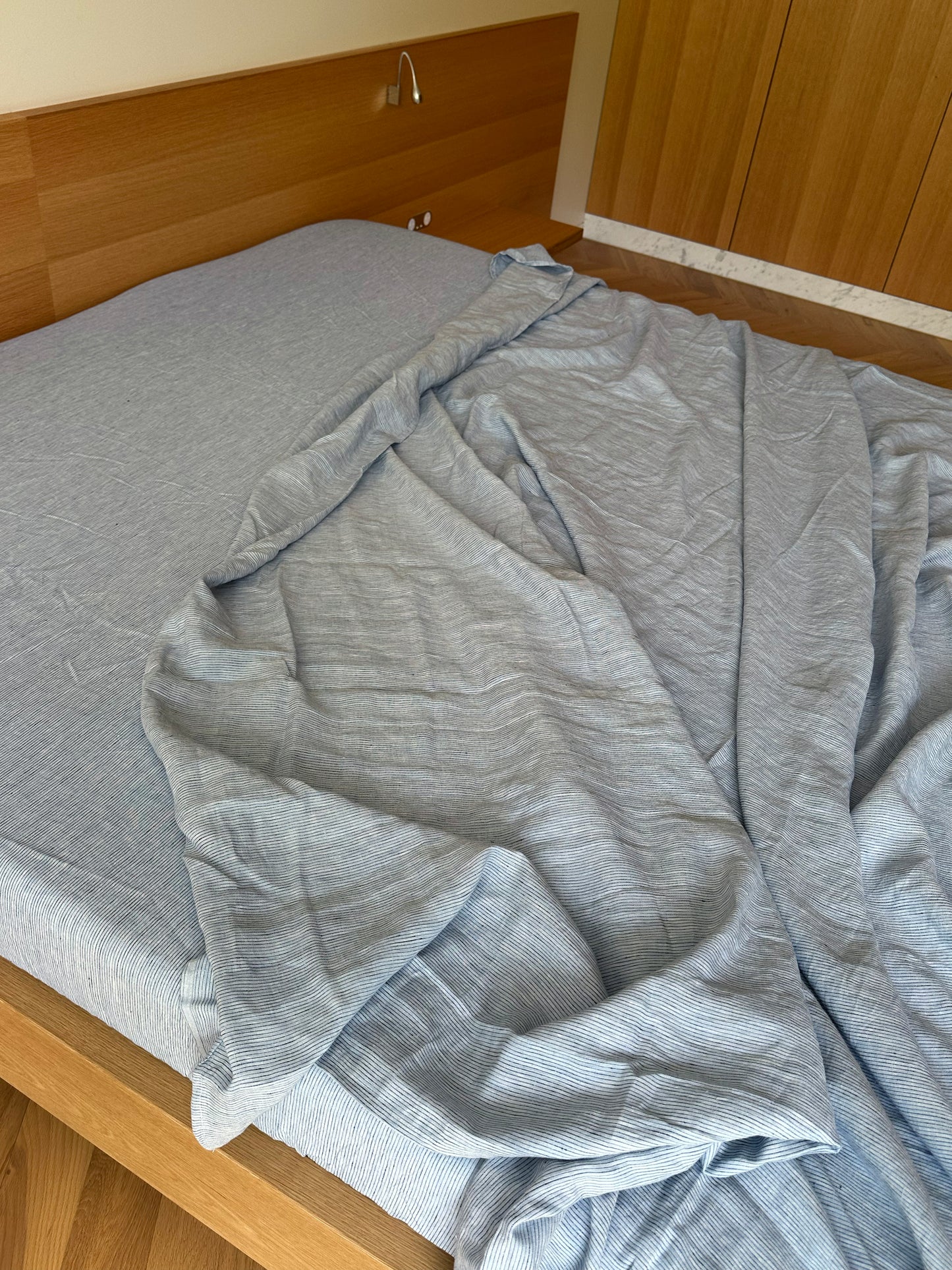 A wooden bed made up with Sheets - Blue Stripe—a light blue fitted sheet and a casually draped, untucked flat sheet—in a bright room featuring wooden floors and cabinets.