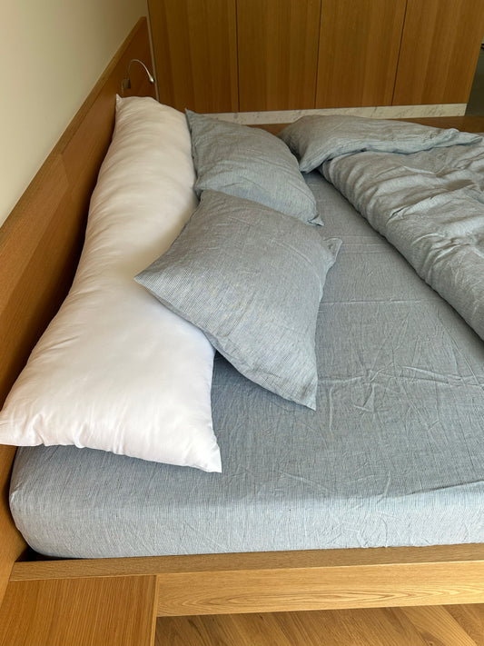 A neatly made wooden bed with hypo-allergenic linen sheets, two gray pillows, a long white body pillow, and a Blue Stripe duvet cover folded at the side, in a room with wooden flooring and a wardrobe.