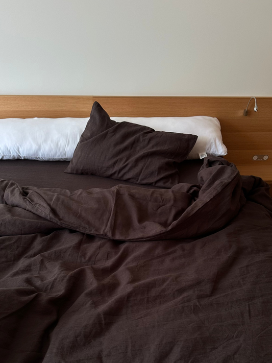 A bed with a wooden headboard and attached nightstand features dark brown bedding, an unmade duvet, and a large white pillow in the Pillow slip set - Bark. A brown pillow is also placed at the head of the bed.