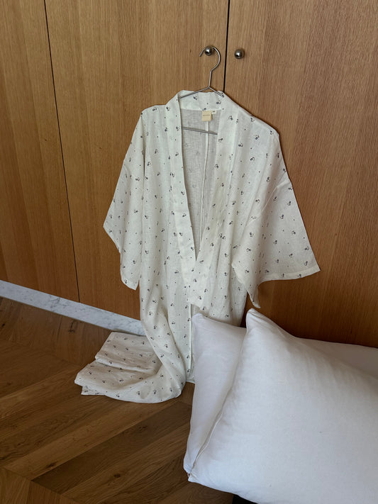 A vintage floral, light-colored full-length 02 robe with a subtle pattern and linen waist tie hangs on a hook by a wooden closet; a white pillow rests on the floor nearby.