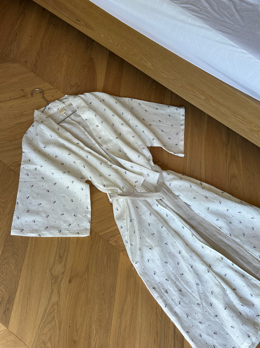 The 02 robe - vintage floral is a full-length, wide-sleeved robe with a subtle small pattern, linen waist tie, and hanger attached to the collar, displayed flat on a wooden floor next to a bed with a white sheet.