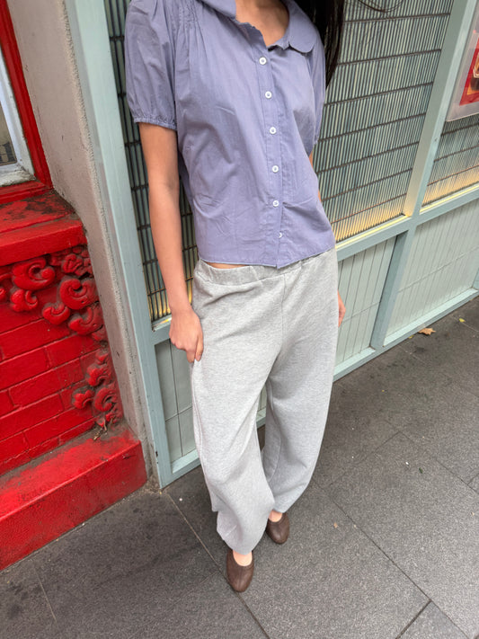 A cropped view of a woman on a street wearing the Petal Top - Thistle by Deiji Studios, a pale periwinkle petal-collar blouse with front buttons and puff sleeves, with grey marle trousers.