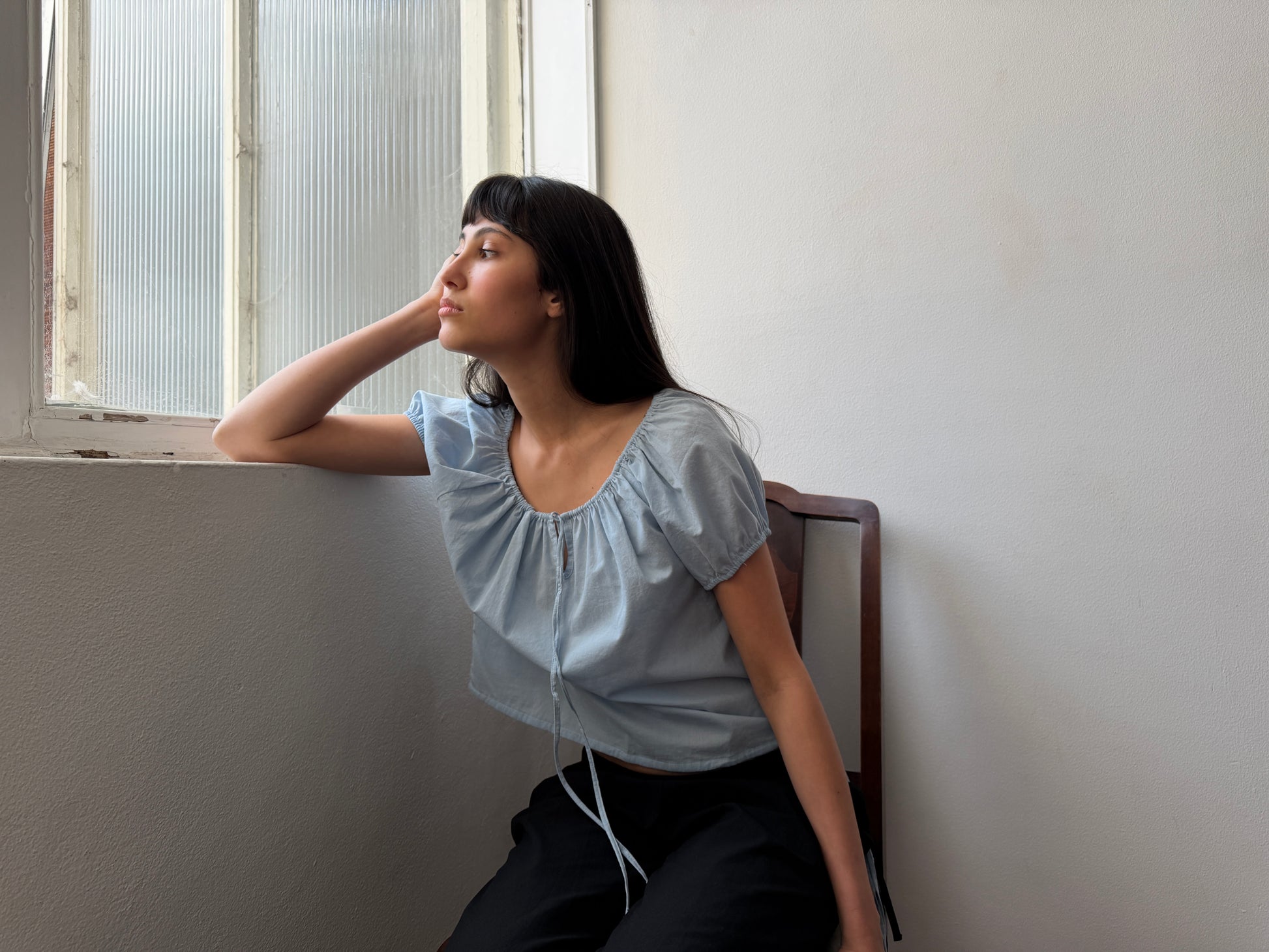 A woman with long dark hair sits on a chair by a window, resting her elbow on the windowsill and gazing outside thoughtfully. She wears the Tie Top - Blue by Deiji Studios, paired with black pants.