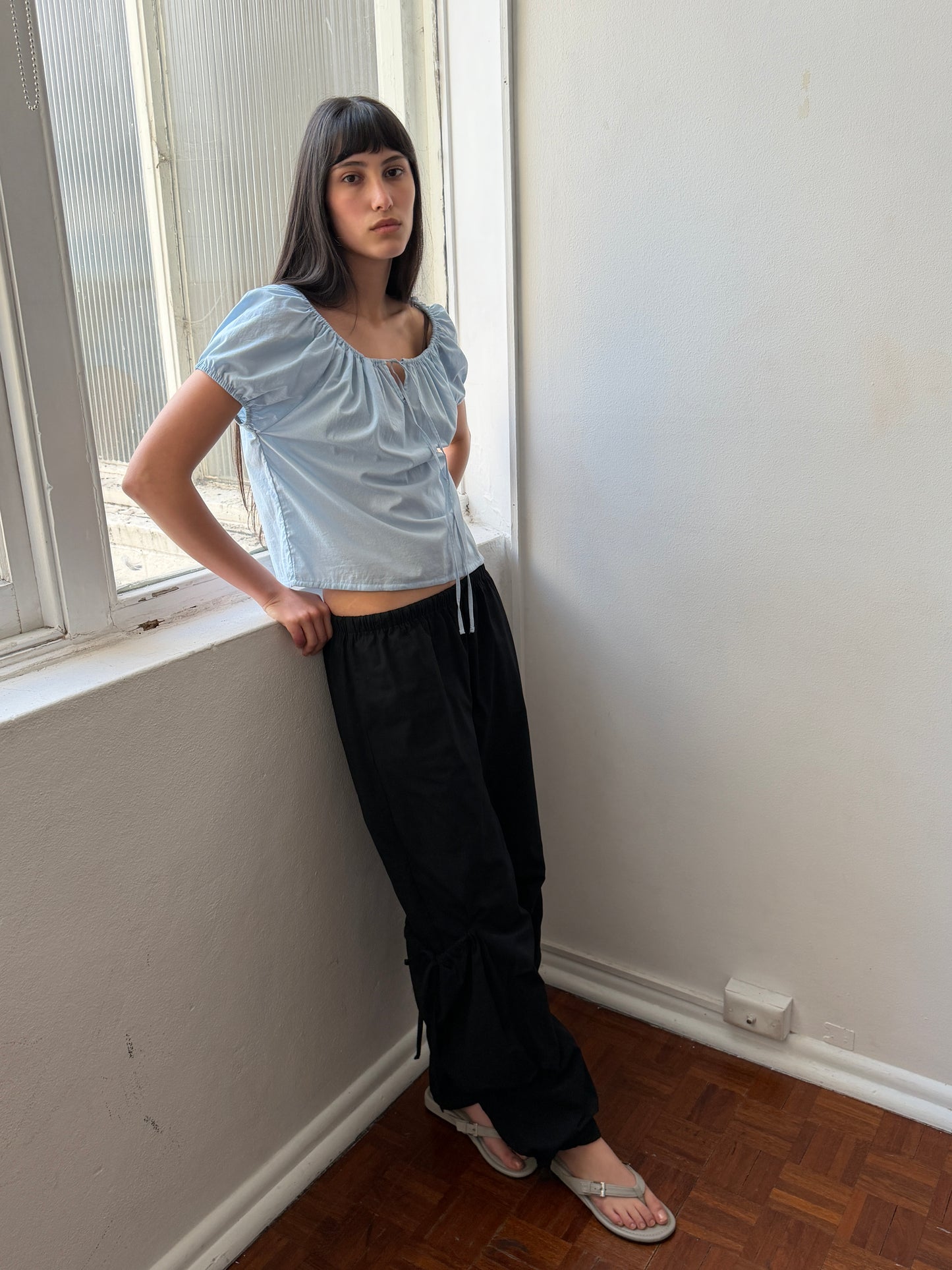 A person with straight dark hair, dressed in a light blue top and the Drawcord Pocket Pant - Black by Deiji Studios, paired with flip-flops, stands by a large window in a room featuring white walls and a wooden floor.
