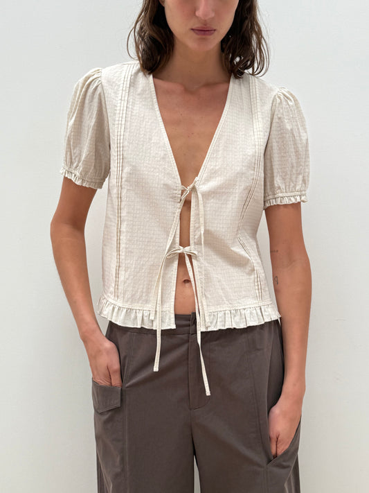 A person wears the Pintuck Top - Bone Check, an organic cotton, cream-toned pintuck top with short sleeves, tie-front, deep V-neck, and ruffled hem, paired with high-waisted loose-fitting gray pants from Deiji Studios against a white background.