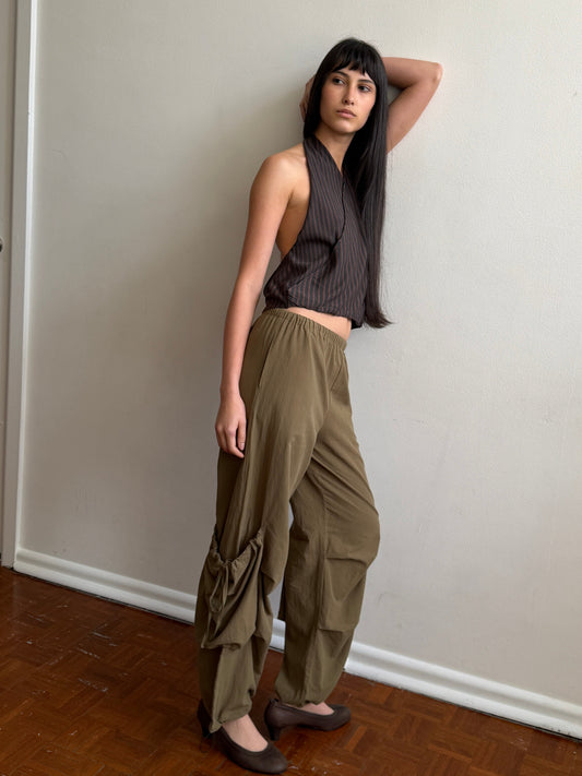 A woman with dark hair and a fringe leans against a wall in a room wearing a brown striped halter top and the Drawcord Pocket Pant - Khaki by Deiji Studios, khaki wide-leg trousers with drawcord-gathered lower legs, and flats.