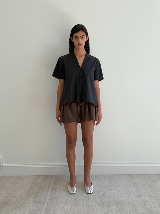 A woman stands against a plain, light wall, wearing the Sails Top in Black Stripe with loose brown shorts and silver flats. She looks directly at the camera, arms relaxed by her sides.