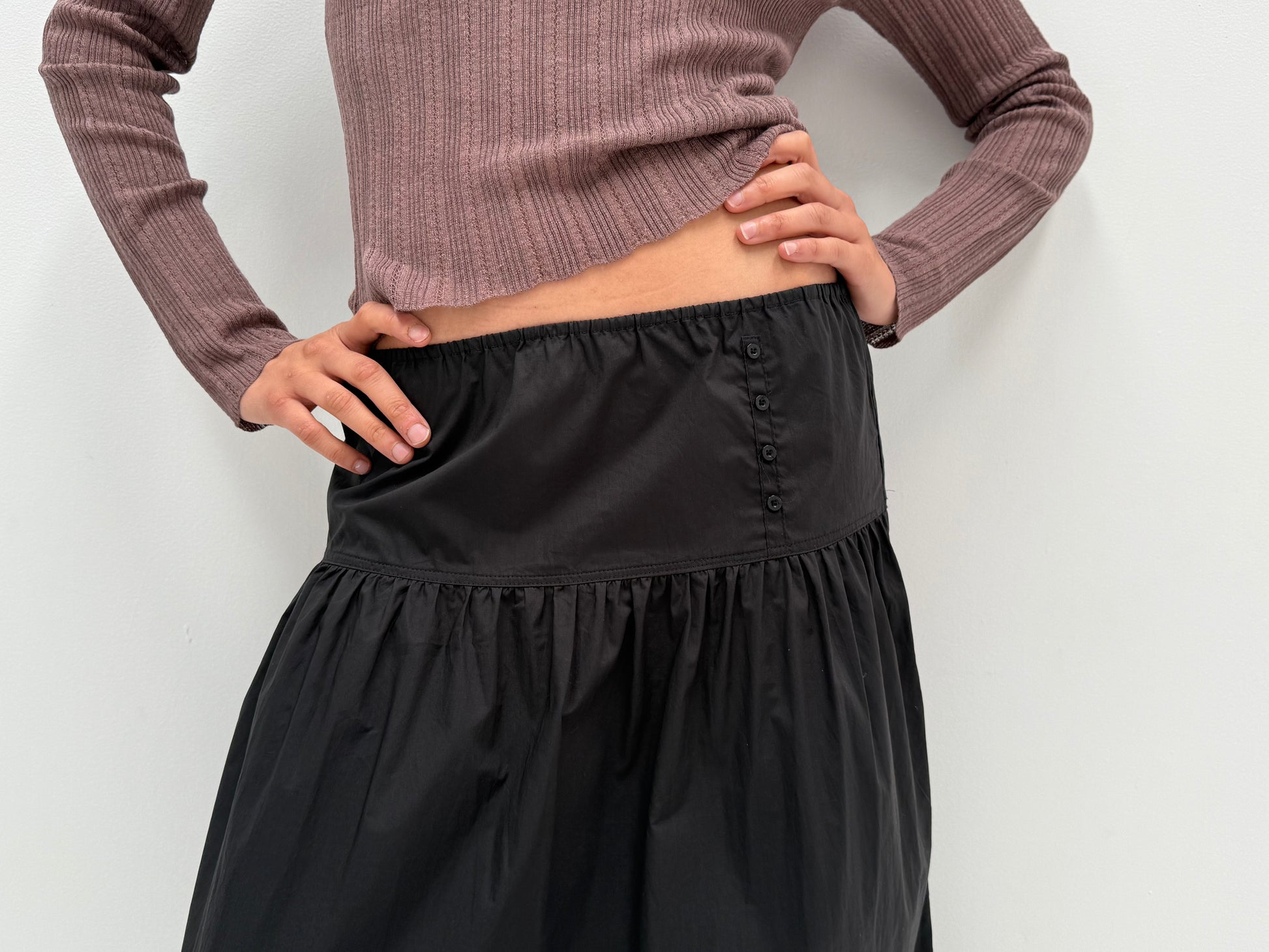 A person stands with hands on hips against a plain white background, wearing a ribbed, long-sleeve mauve top and the Button Skirt - Black.