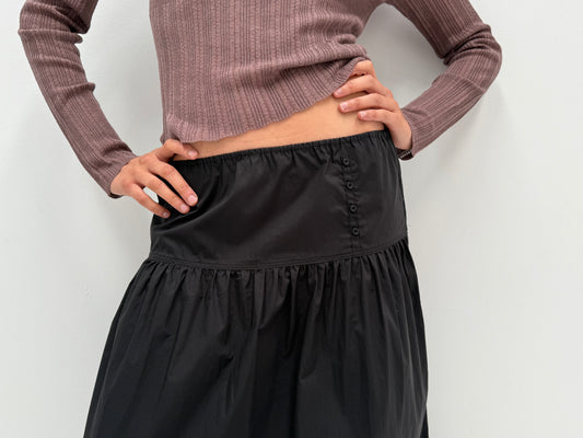 A person stands with hands on hips against a plain white background, wearing a ribbed, long-sleeve mauve top and the Button Skirt - Black.