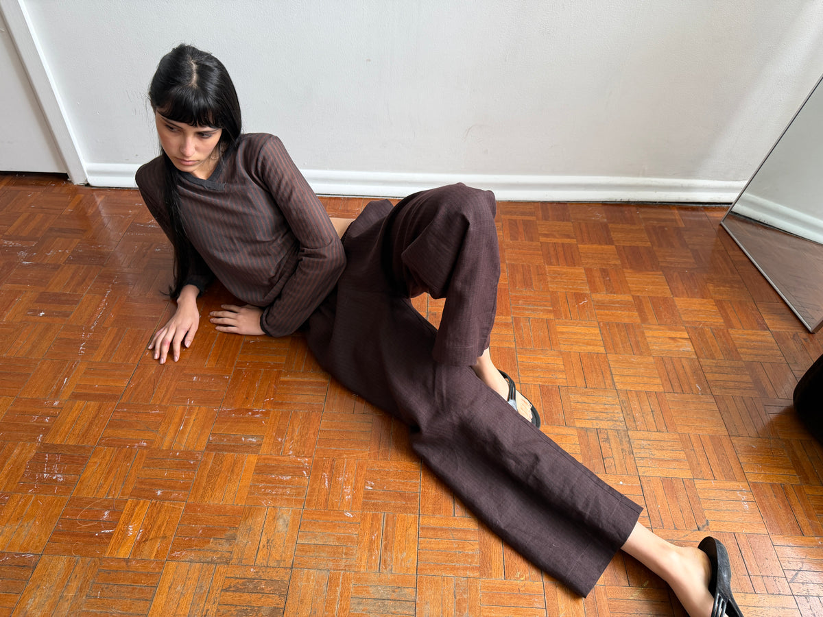A woman with dark hair and a fringe reclines on a parquet floor propped on one arm, wearing the Linen Ease Trouser - Brown Black Stripe by Deiji Studios, dark brown fine-striped wide-leg linen trousers, with a matching brown striped knit top.