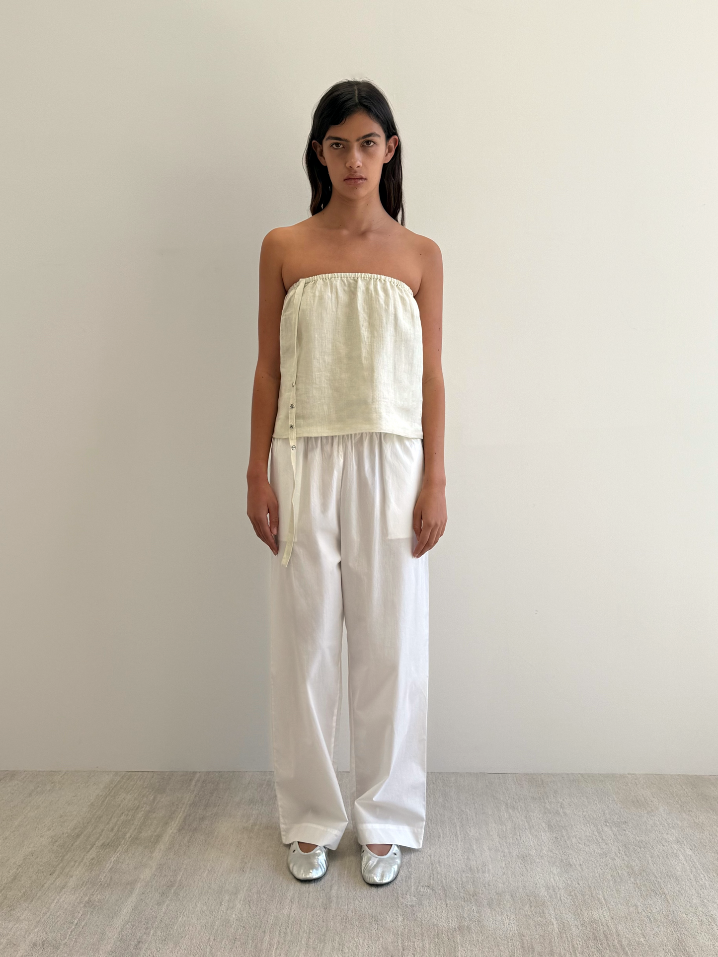 A woman with straight dark hair stands against a white wall, wearing the Button Strap Top in Pastel Pistachio with side ties, loose white pants, and silver shoes on a light-colored floor.