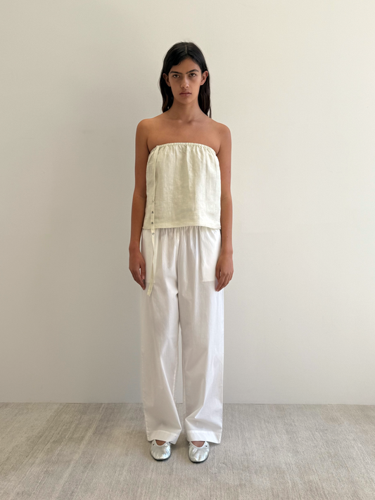 A woman with straight dark hair stands against a white wall, wearing the Button Strap Top in Pastel Pistachio with side ties, loose white pants, and silver shoes on a light-colored floor.