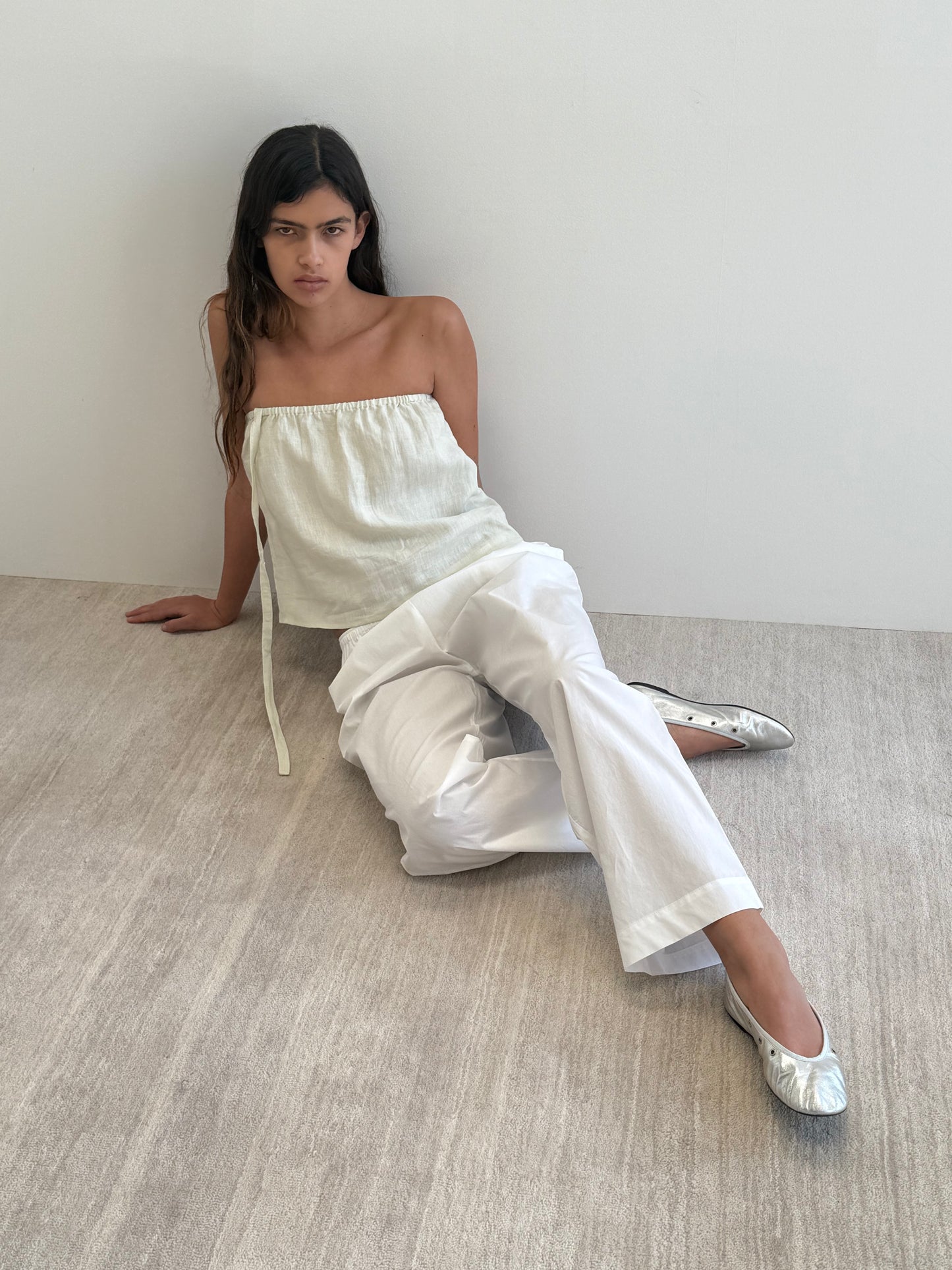 A woman with long dark hair sits on the floor against a white wall in a minimal, neutral setting, wearing the Button Strap Top in Pastel Pistachio, loose white pants, and silver flat shoes.