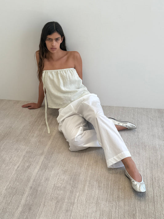 A woman with long dark hair sits on the floor against a white wall in a minimal, neutral setting, wearing the Button Strap Top in Pastel Pistachio, loose white pants, and silver flat shoes.
