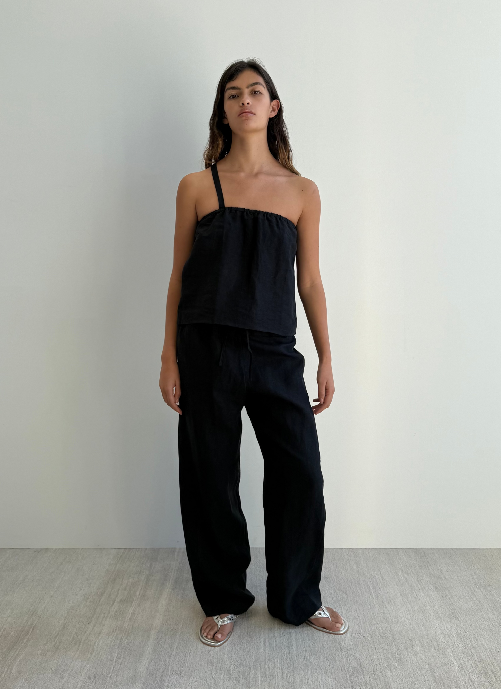 A woman with long dark hair and a neutral expression stands against a white wall wearing Deiji Studios’ black Button Strap Top in organic French linen, matching wide-leg pants, and silver sandals.