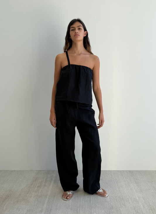 A woman with long dark hair and a neutral expression stands against a white wall wearing Deiji Studios’ black Button Strap Top in organic French linen, matching wide-leg pants, and silver sandals.