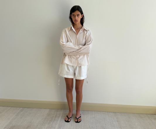 A woman with straight dark hair and a neutral expression stands indoors against a light wall, wearing the Deiji Studios Room Shirt - Dusty Stripe, loose white shorts, and black sandals. Her arms are relaxed by her sides.
