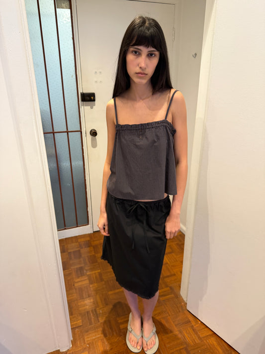 A woman with dark hair and a fringe stands in a hallway wearing the One Frill Skirt - Black by Deiji Studios, a black knee-length skirt with a drawstring waist and frill hem, with a grey striped cami top and flip-flops.