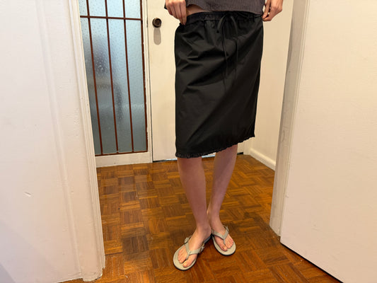 A cropped view of the One Frill Skirt - Black by Deiji Studios, a black knee-length skirt with a drawstring waist and single frill hem, worn with flip-flops in a hallway.