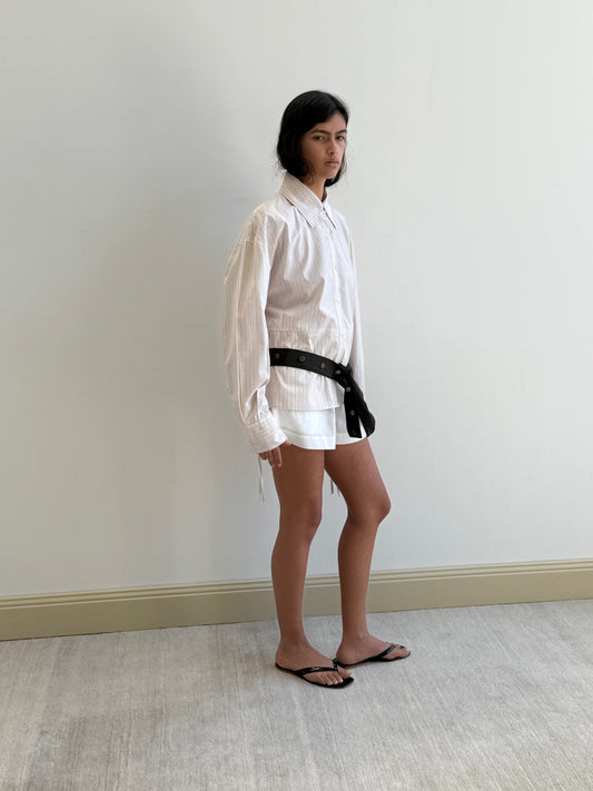 A person with long dark hair and a neutral expression stands indoors by a white wall, wearing the Deiji Studios Room Shirt - Dusty Stripe, paired with white shorts, a black belt, and black sandals.